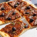 Sliced Pissaladière on parchment paper next to a glass of juice and a bowl of kalamata olives.