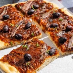 Sliced Pissaladière on parchment paper next to a glass of juice and a bowl of kalamata olives.
