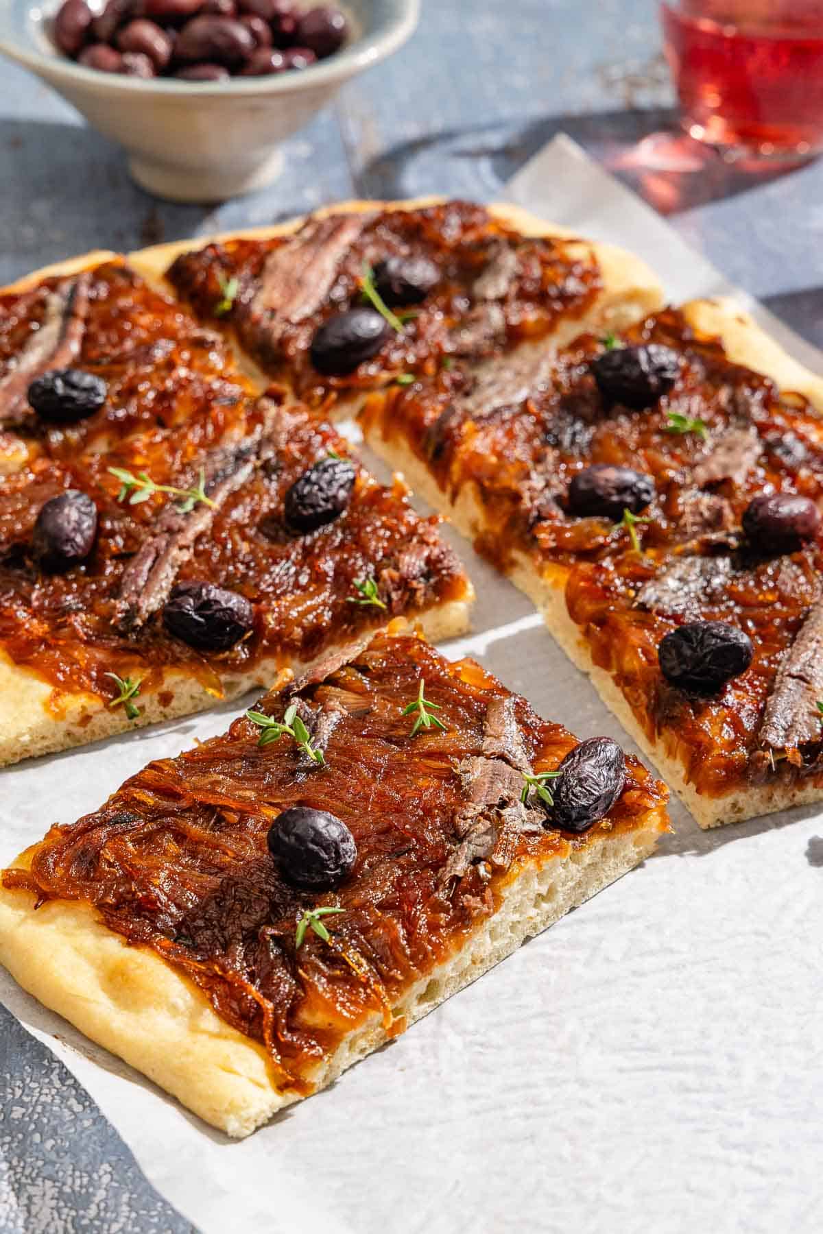 Sliced Pissaladière on parchment paper next to a glass of juice and a bowl of kalamata olives.