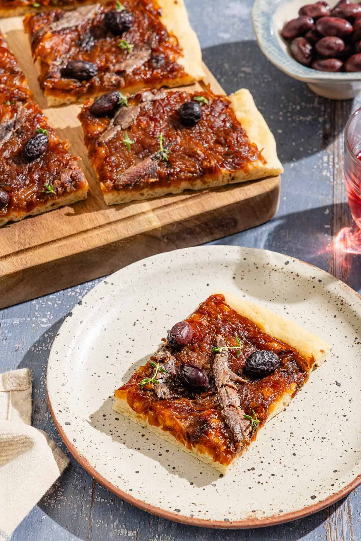 A piece of Pissaladière on a plate, with the rest of the Pissaladière on a wooden cutting board and a bowl of kalamata olives in the background.