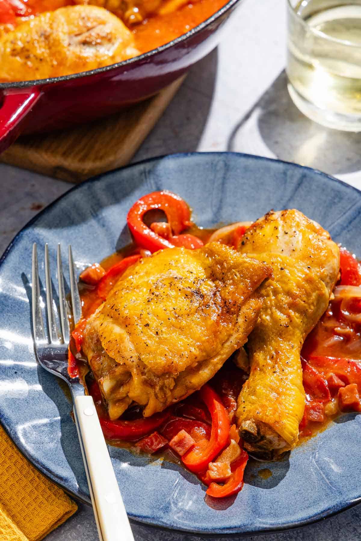 A serving of chicken and peppers on a plate with a fork next to chicken and peppers in a skillet and a glass of white wine.