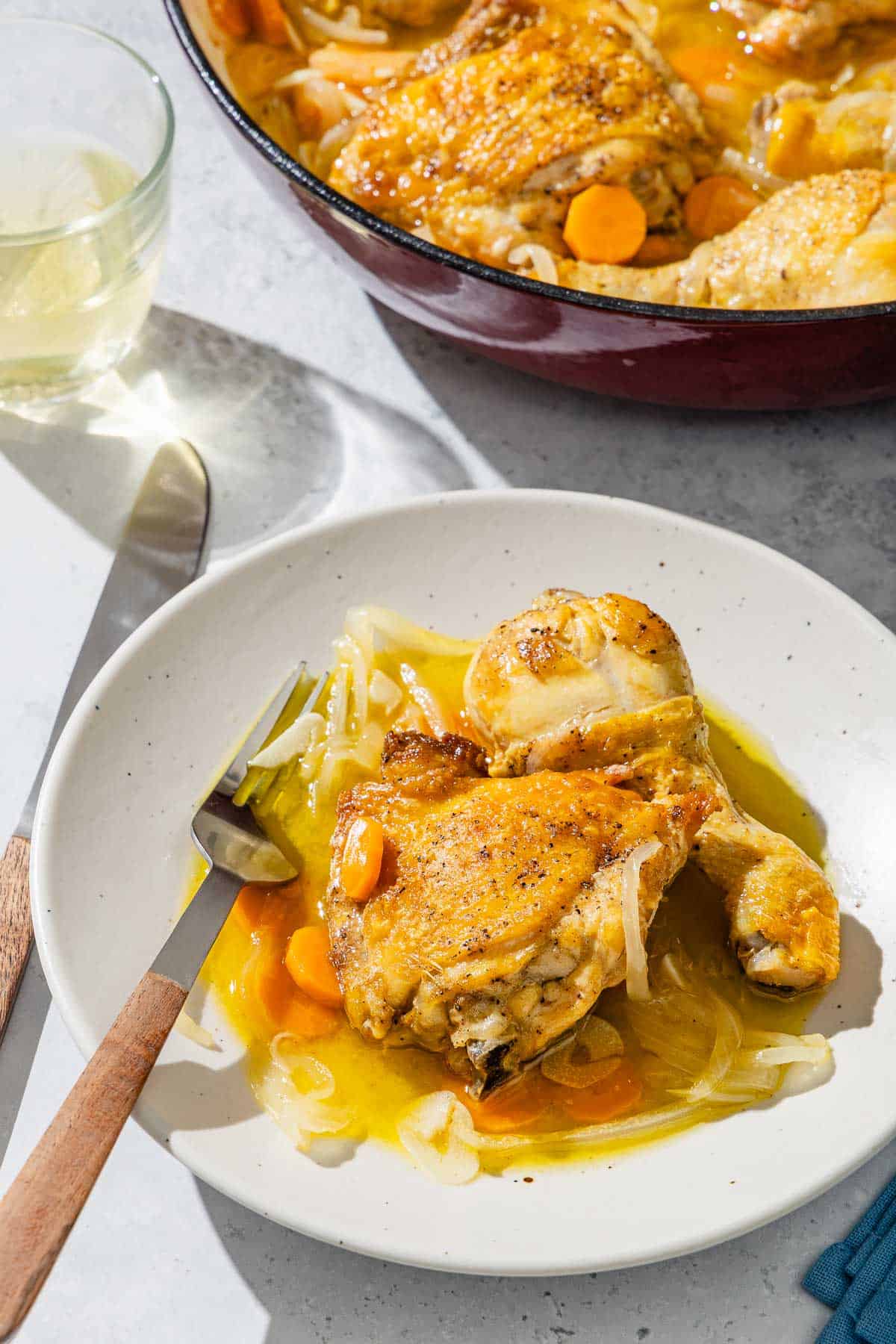 A serving of escabeche de pollo on a plate with a fork. Next to this is a knife, a glass of white wine, and the rest of the escabeche de pollo in a skillet.