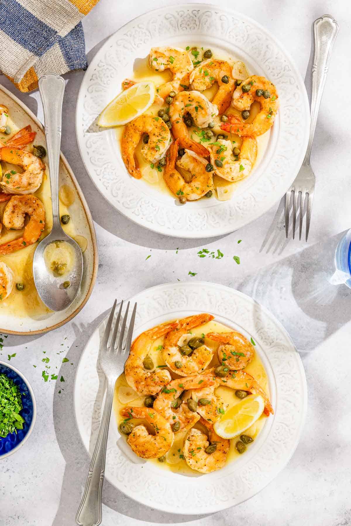 Two servings of shrimp piccata with lemon wedges on plates with forks. Next to this is the rest of the shrimp piccata on a platter with a serving spoon and a bowl of chopped parsley.