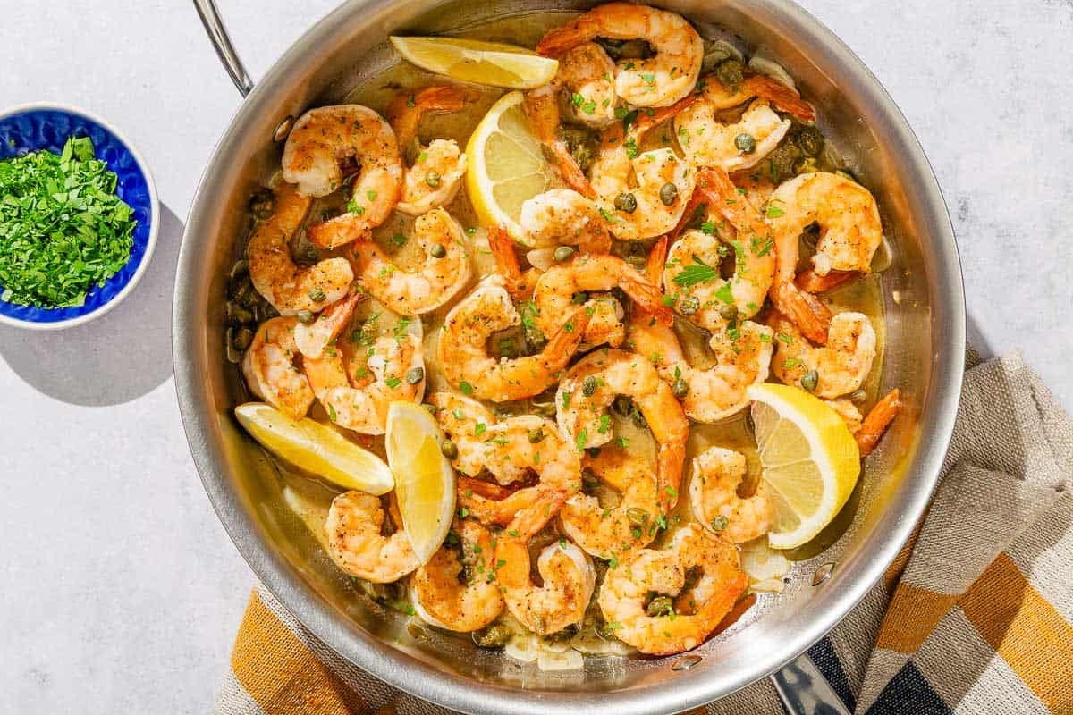 The shrimp piccata with lemon wedges in a skillet next to a small bowl of chopped parsley.