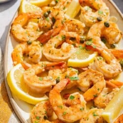 Shrimp piccata with lemon wedges on a platter next to small bowls of chopped parsley and capers.