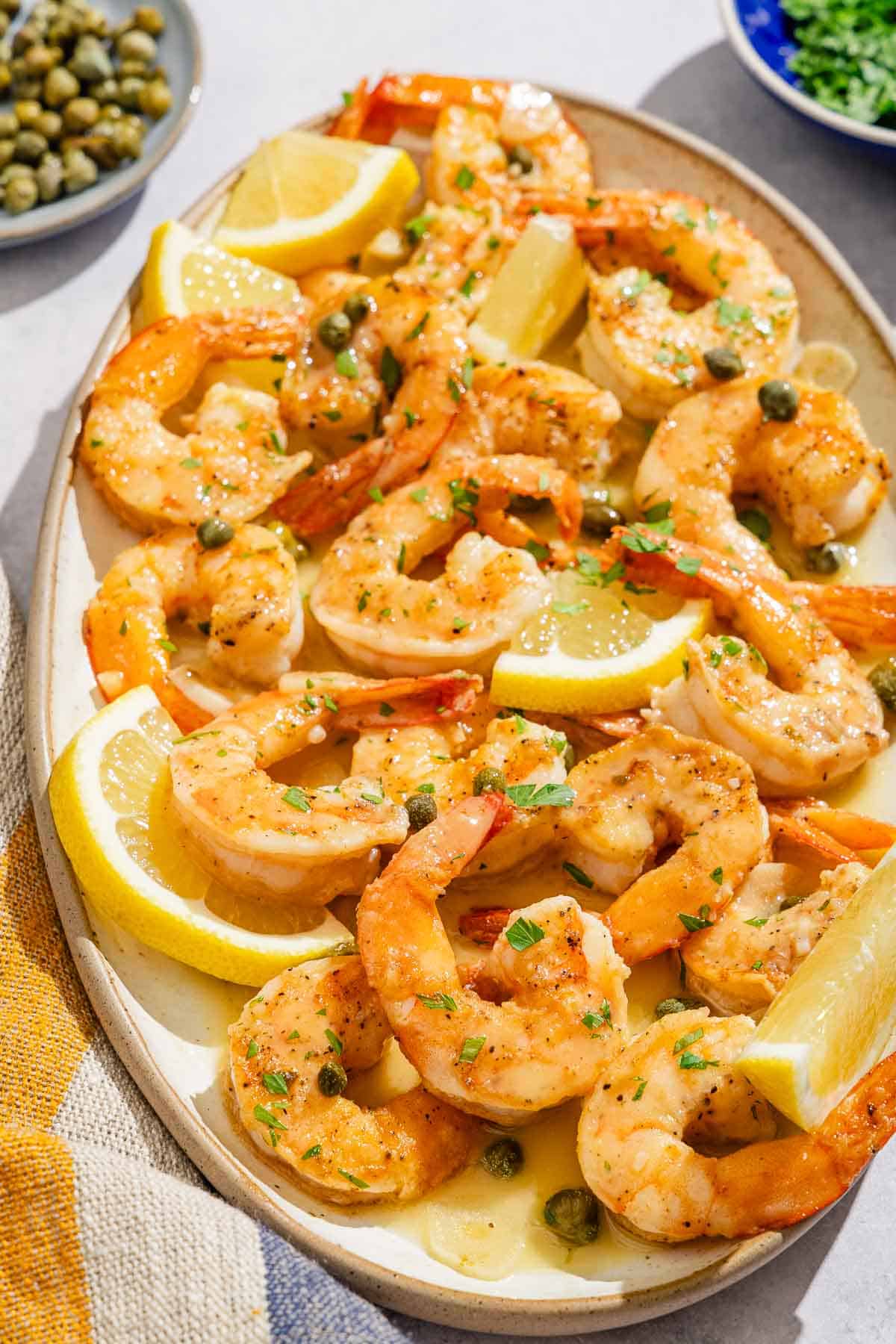 Shrimp piccata with lemon wedges on a platter next to small bowls of chopped parsley and capers.