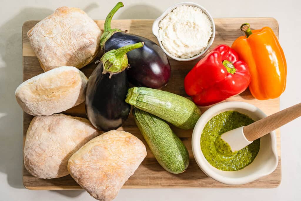 Ingredients for the roasted vegetable sandwich recipe including eggplant, zucchini, bell peppers, olive oil, salt, black pepper, ciabatta rolls, ricotta cheese, and basil pesto.