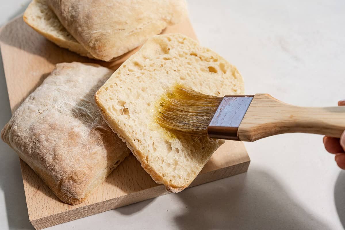 A close up of 4 ciabatta halves on a cutting board, one being brushed with olive oil.