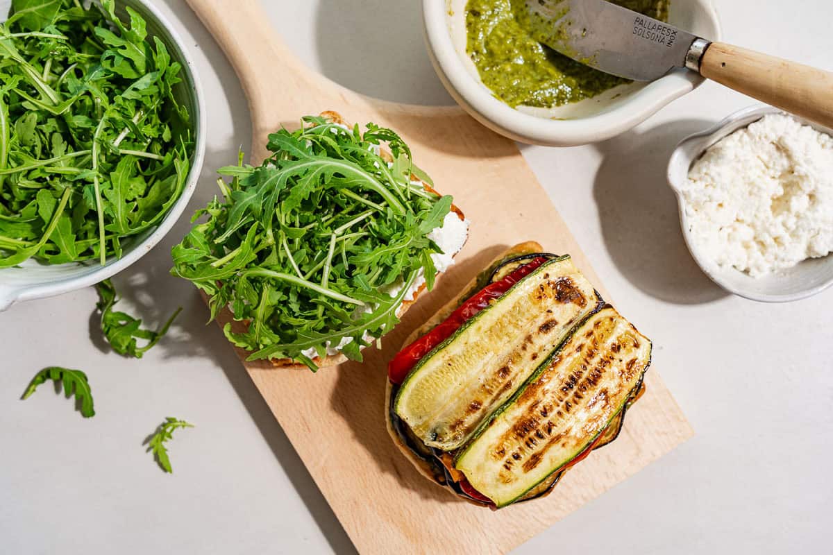 An overhead photo of two toasted ciabatta bread halves on a wooden cutting board. One half topped with basil pesto and roasted vegetables, and one half topped with ricotta and arugula. Next to these are bowls of arugula, basil pesto with a knife, and ricotta.
