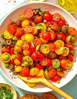 Cherry tomato salad in a serving bowl with a wooden spoon. Next to this is a bowl of chopped parsley, a bottle of olive oil, a bowl of Aleppo pepper and two slices of toasted bread.