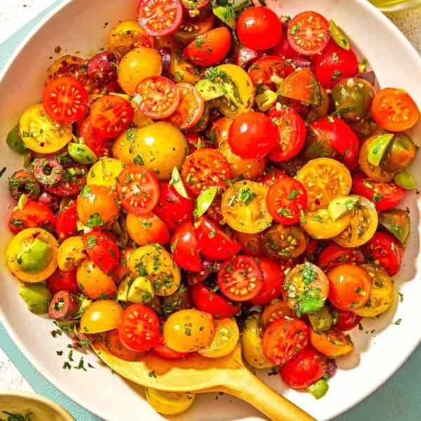 Cherry tomato salad in a serving bowl with a wooden spoon. Next to this is a bowl of chopped parsley, a bottle of olive oil, a bowl of Aleppo pepper and two slices of toasted bread.