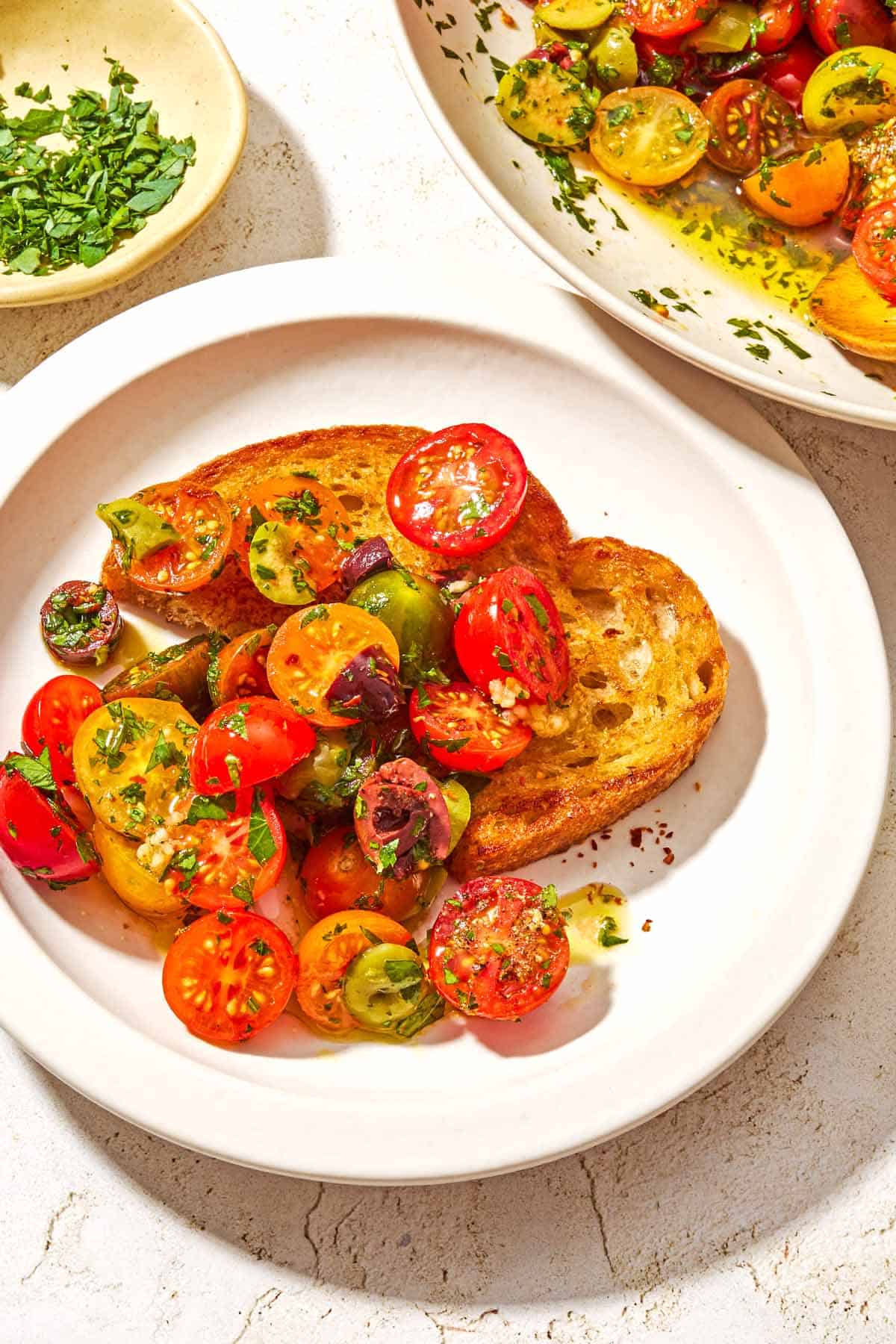 A serving of cherry tomato salad on a plate with a piece of toasted bread. Next to this is the rest of the salad in a serving bowl, and a small bowl of chopped parsley.