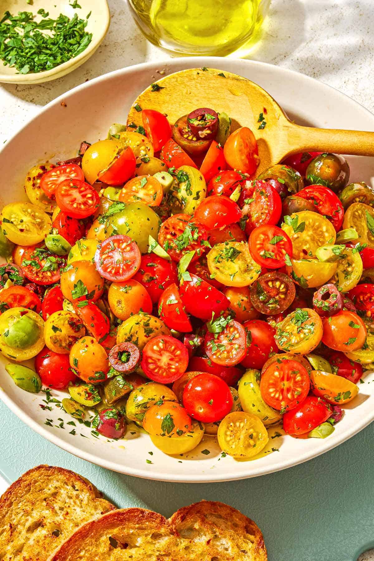 Cherry tomato salad in a serving bowl with a wooden spoon. Next to this is a bowl of chopped parsley, a bottle of olive oil, and two slices of toasted bread.