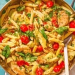 Chicken pasta in a tall-sided skillet with a spoon. Next to this is a kitchen towel and a small bowl of chopped parsley.