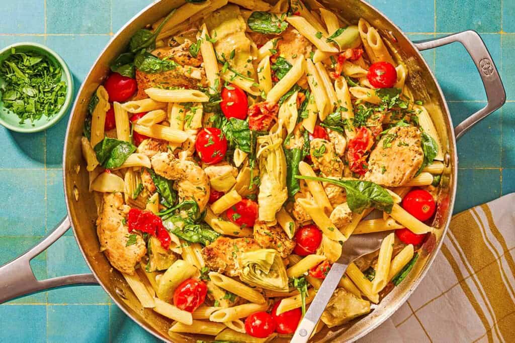 Chicken pasta in a tall-sided skillet with a spoon. Next to this is a kitchen towel and a small bowl of chopped parsley.