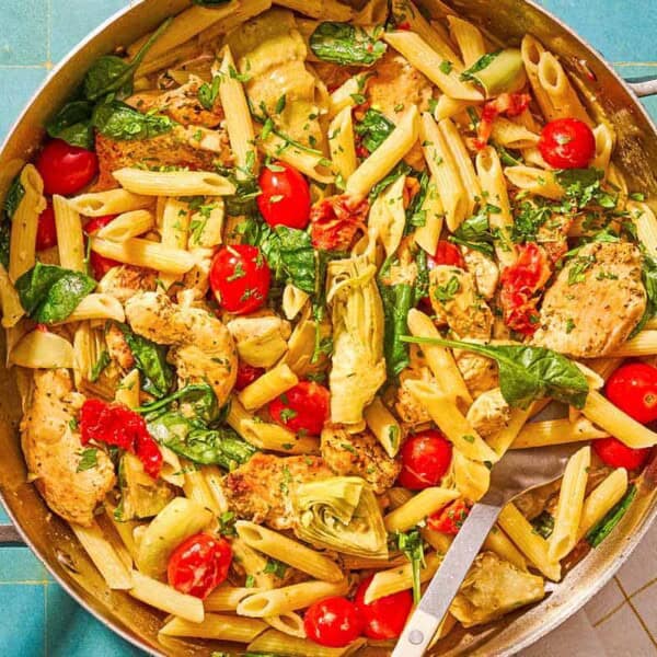 Chicken pasta in a tall-sided skillet with a spoon. Next to this is a kitchen towel and a small bowl of chopped parsley.