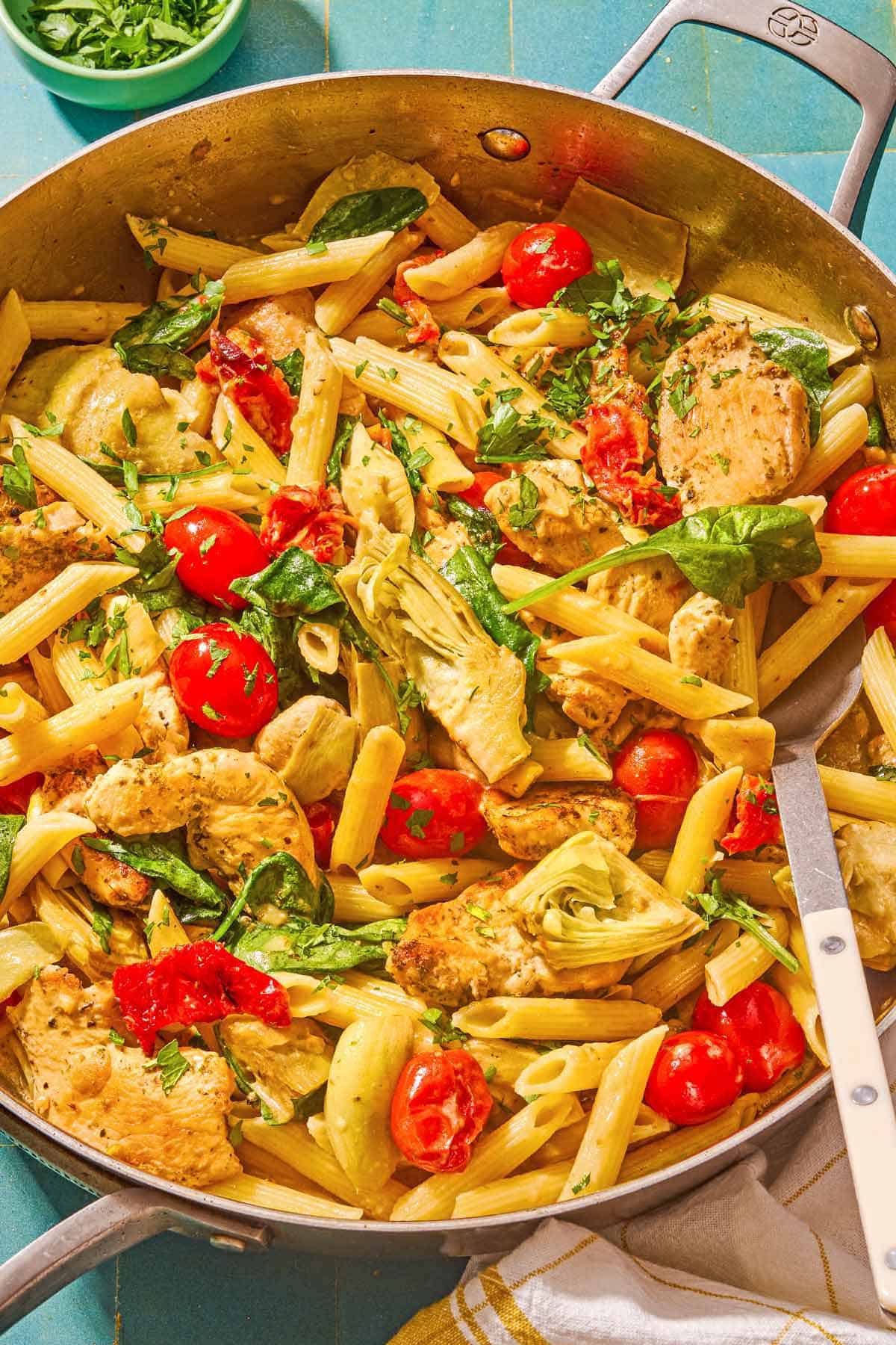 Chicken pasta in a tall-sided skillet with a spoon. Next to this is a kitchen towel and a small bowl of chopped parsley.