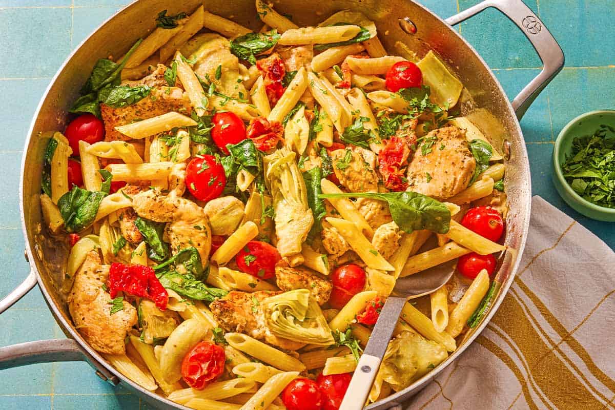 Chicken pasta in a tall-sided skillet with a spoon. Next to this is a kitchen towel and a small bowl of chopped parsley.