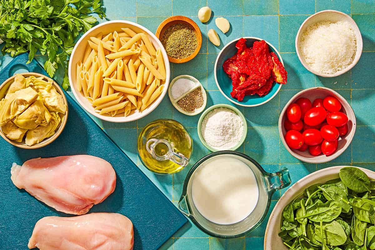 Ingredients for the chicken pasta recipe including chicken, penne pasta, salt, black pepper, dried oregano, olive oil garlic, sun dried tomatoes, cherry tomatoes, artichoke hearts, milk, flour, grated parmesan, baby spinach and parsley.