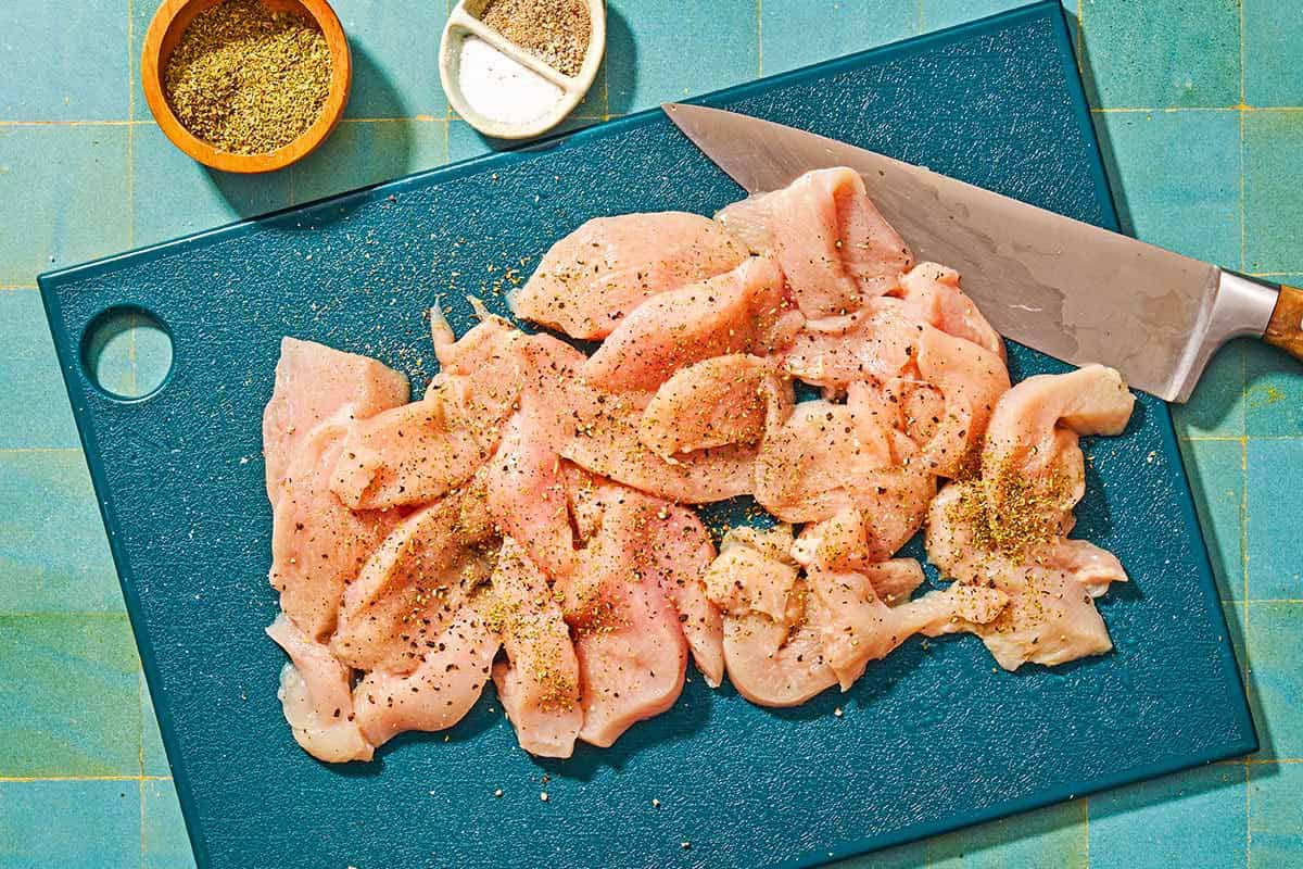 The uncooked chicken season with salt, black pepper and dried oregano cut into pieces on a cutting board with a knife. Next to this are bowls of dried oregano, salt and black pepper.