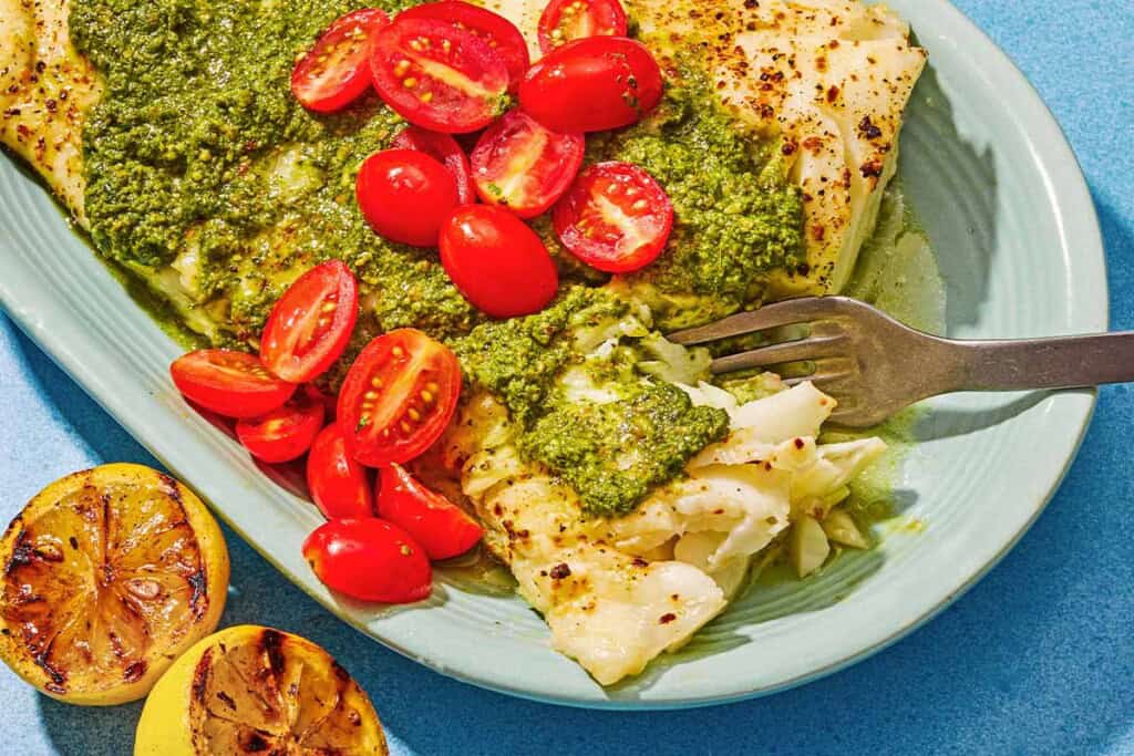 Chilean sea bass topped with pesto and grape tomatoes on a platter with a fork. Next to this is two grilled lemon halves.