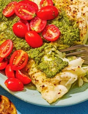 Chilean sea bass topped with pesto and grape tomatoes on a platter with a fork. Next to this is two grilled lemon halves.