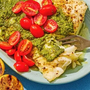 Chilean sea bass topped with pesto and grape tomatoes on a platter with a fork. Next to this is two grilled lemon halves.