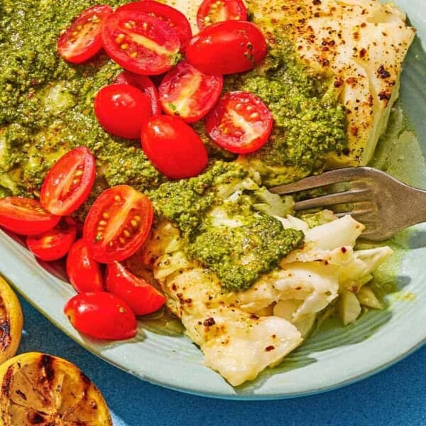 Chilean sea bass topped with pesto and grape tomatoes on a platter with a fork. Next to this is two grilled lemon halves.