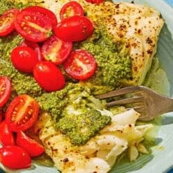 A close up of Chilean sea bass topped with pesto and grape tomatoes on a platter along with a grilled lemon half and a fork.