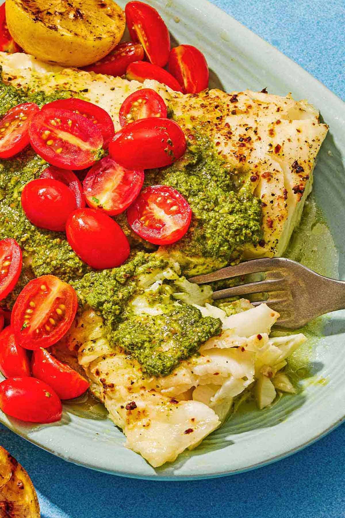 A close up of Chilean sea bass topped with pesto and grape tomatoes on a platter along with a grilled lemon half and a fork.