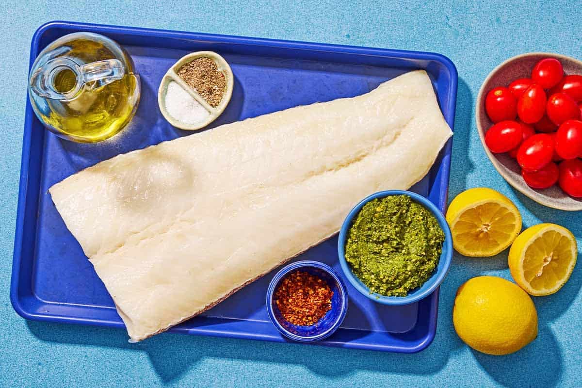 The ingredients for Chilean Sea Bass including Chilean sea bass, olive oil, salt, black pepper, lemons, pesto, and grape tomatoes.