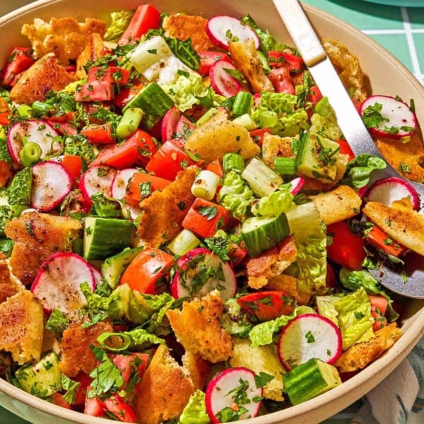 Fattoush in a serving bowl with a spoon. Next to this is a small bowl of the dressing and sumac, a cloth napkin, and a plate.