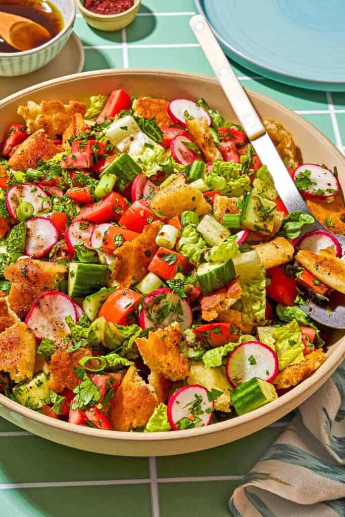 Fattoush in a serving bowl with a spoon. Next to this is a small bowl of the dressing and sumac, a cloth napkin, and a plate.