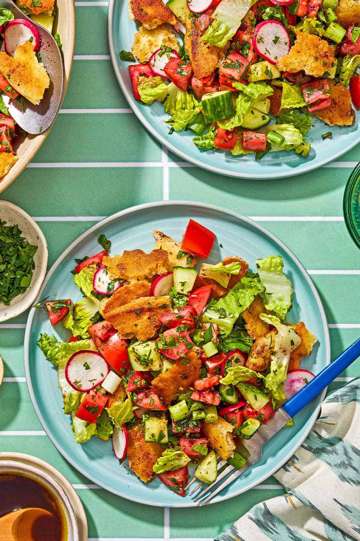 A serving of fattoush salad on a plate with a fork. Surrounding this is another plate of the fattoush, small bowls of chopped parsley and the salad dressing, and a cloth napkin.
