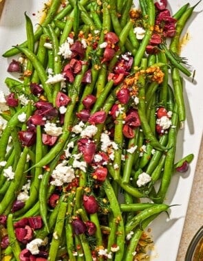 Green bean salad on a platter next to bowls of feta and red pepper flakes, and the dressing for the salad in a measuring cup.