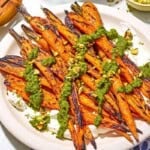 Grilled carrots with labneh topped with zhoug and chopped pistachios on a platter next to a kitchen towel and wooden serving utensils.