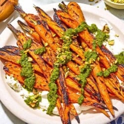 Grilled carrots with labneh topped with zhoug and chopped pistachios on a platter next to a kitchen towel and wooden serving utensils.