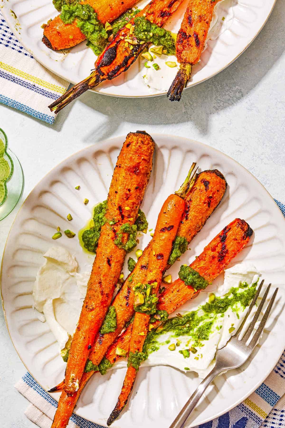 A serving of grilled carrots with labneh topped with zhoug and chopped pistachios on a plate. Next to this is second serving on a plate and 2 cloth napkins.