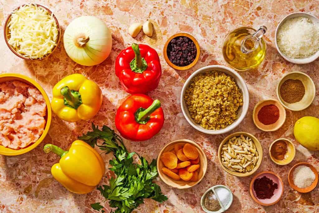 Ingredients for grilled stuffed peppers including bell peppers, freekeh, olive oil, onion, garlic, ground chicken, salt, sugar black pepper, cumin, cinnamon, clove powder, paprika, slivered toasted almonds, dried apricots, currants, parsley, grated parmesan cheese, grated cheddar cheese, and lemon.