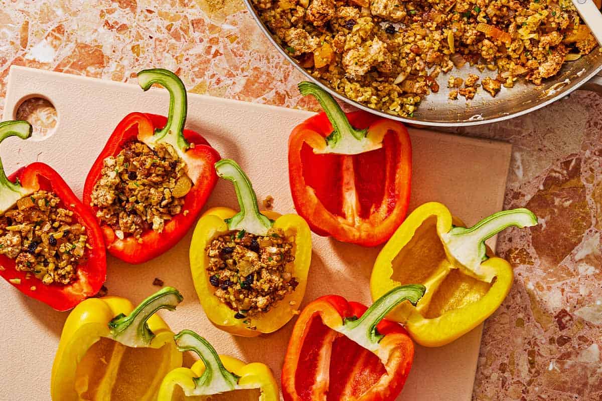 8 bell pepper halves on a cutting board with 3 of them stuffed with the meat mixture. Next to this is the rest of the stuffing in a tall sided skillet.