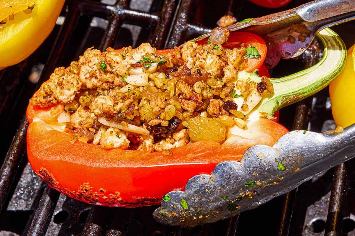 A grilled stuffed pepper on a grill being adjusted with a pair of tongs.