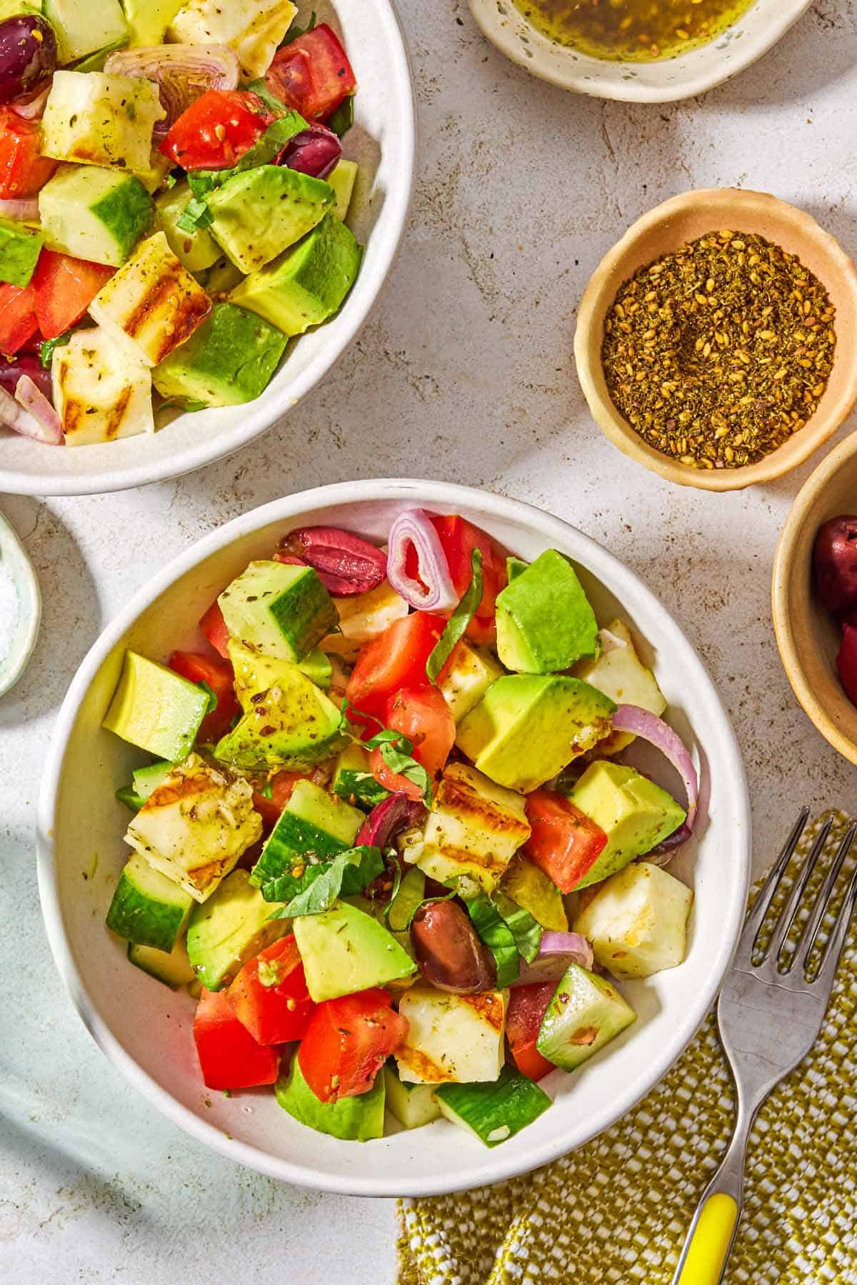 Two bowls of avocado salad surrounded by bowls of the dressing, za'atar, and a fork.