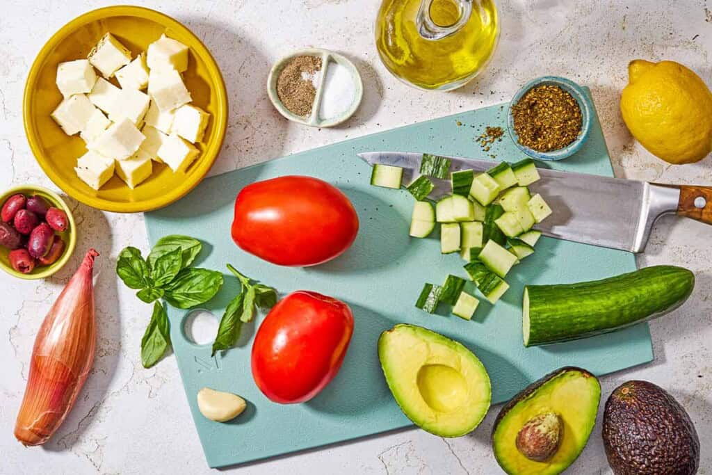 Ingredients for avocado salad including avocados, tomatoes, cucumber, shallot, kalamata olives, basil, halloumi cheese, olive oil, lemon, garlic, za'atar, salt and black pepper.
