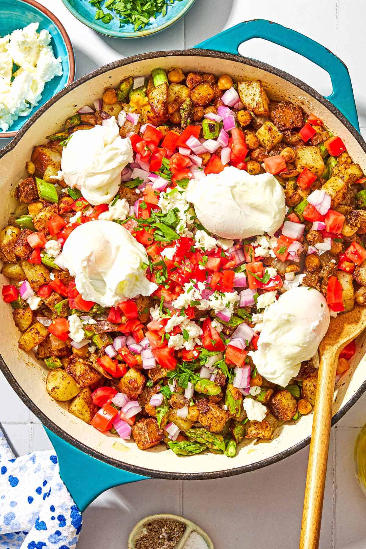 Potato hash with asparagus, chickpeas and feta in a skillet with a wooden spoon. Next to this are bowls of salt, black pepper, feta and chopped parsley.