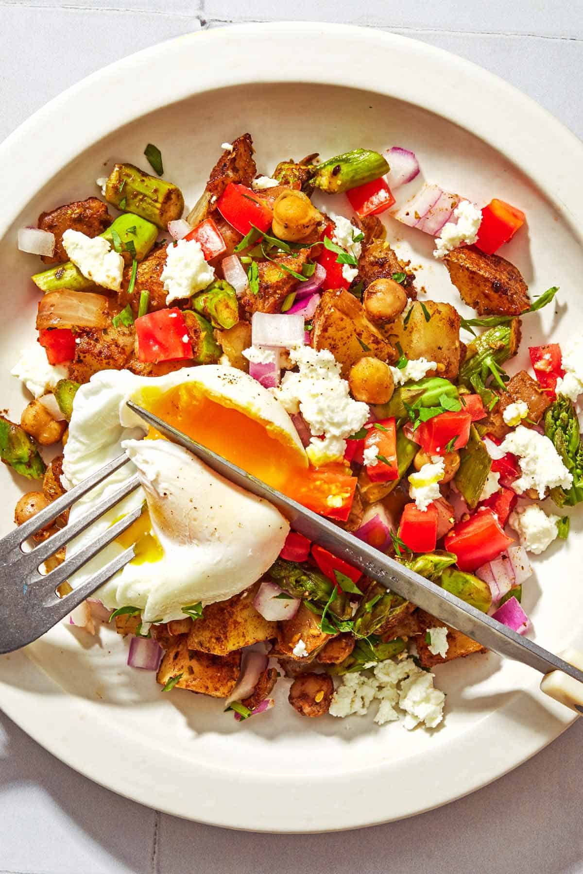 A serving of potato hash with asparagus, chickpeas and feta on a plate with the poached egg on top being cut into with a knife and fork.