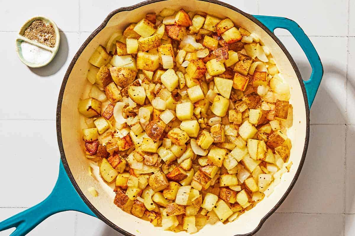 The potatoes, onion and garlic for the potato hash being sauteed in a skillet next to a small bowl with salt and black pepper.