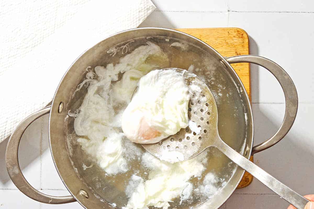 A poached egg being lifted from a saucepan of boiling water with a spoon.