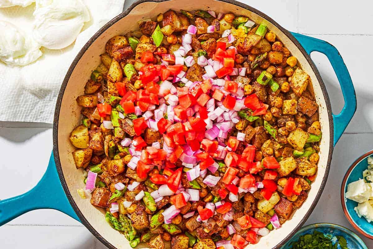The vegetables for the potato hash in a skillet. Next to this are bowls of feta and chopped parsley, and poached eggs resting on a paper towel.