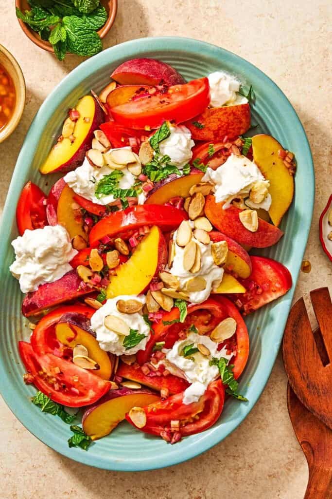 Peach burrata salad on a platter surrounded by a set of serving utensils, and small bowls of the dressing and mint leaves.