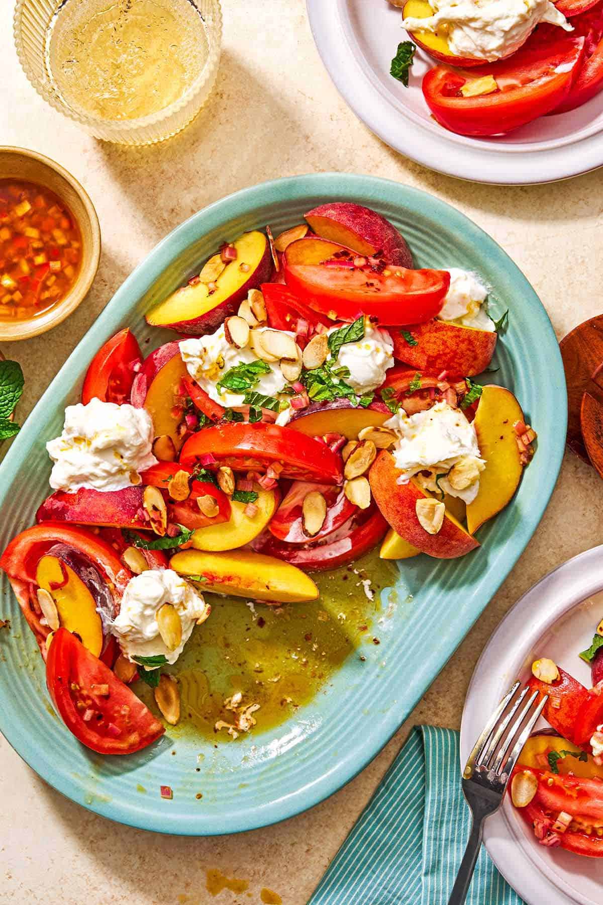 Peach burrata salad on a platter surrounded two servings of the salad on plates, a small bowl of the salad dressing, and a glass of white wine.